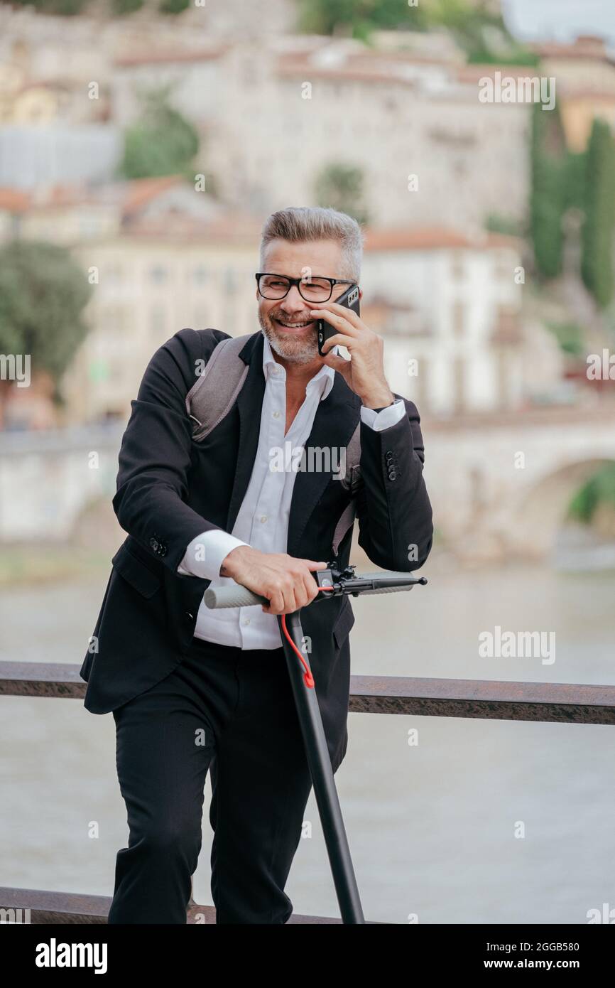 Businessman with elegant suit standing on the electric scooter while talking on the phone. Entrepreneur riding the scooter and answering the phone Stock Photo