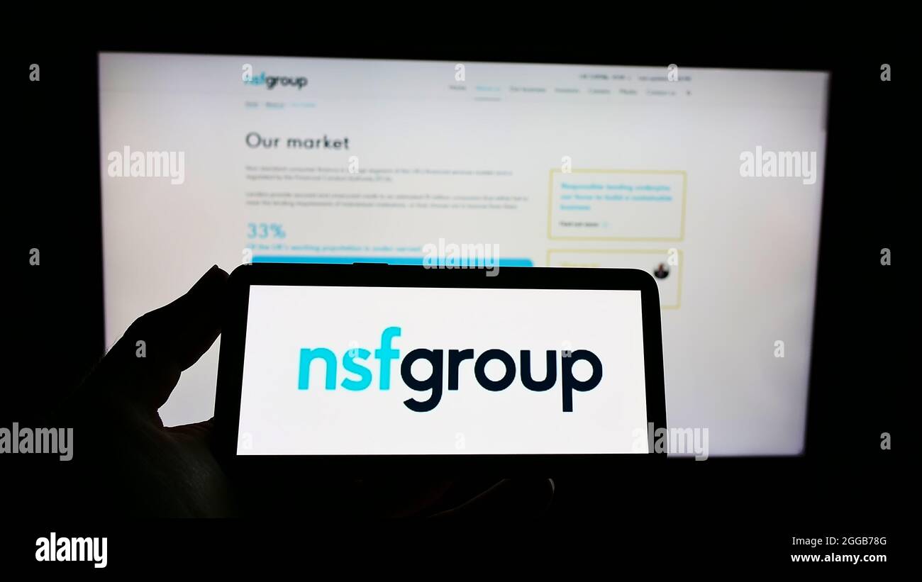 Person holding mobile phone with logo of finance company Non-Standard Finance plc (NSF) on screen in front of web page. Focus on phone display. Stock Photo