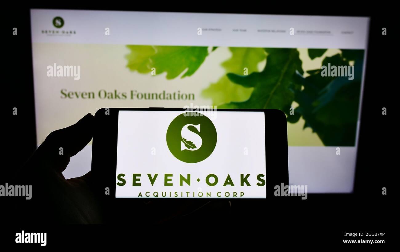 Person holding cellphone with logo of American company Seven Oaks Acquisition Corp. on screen in front of webpage. Focus on phone display. Stock Photo