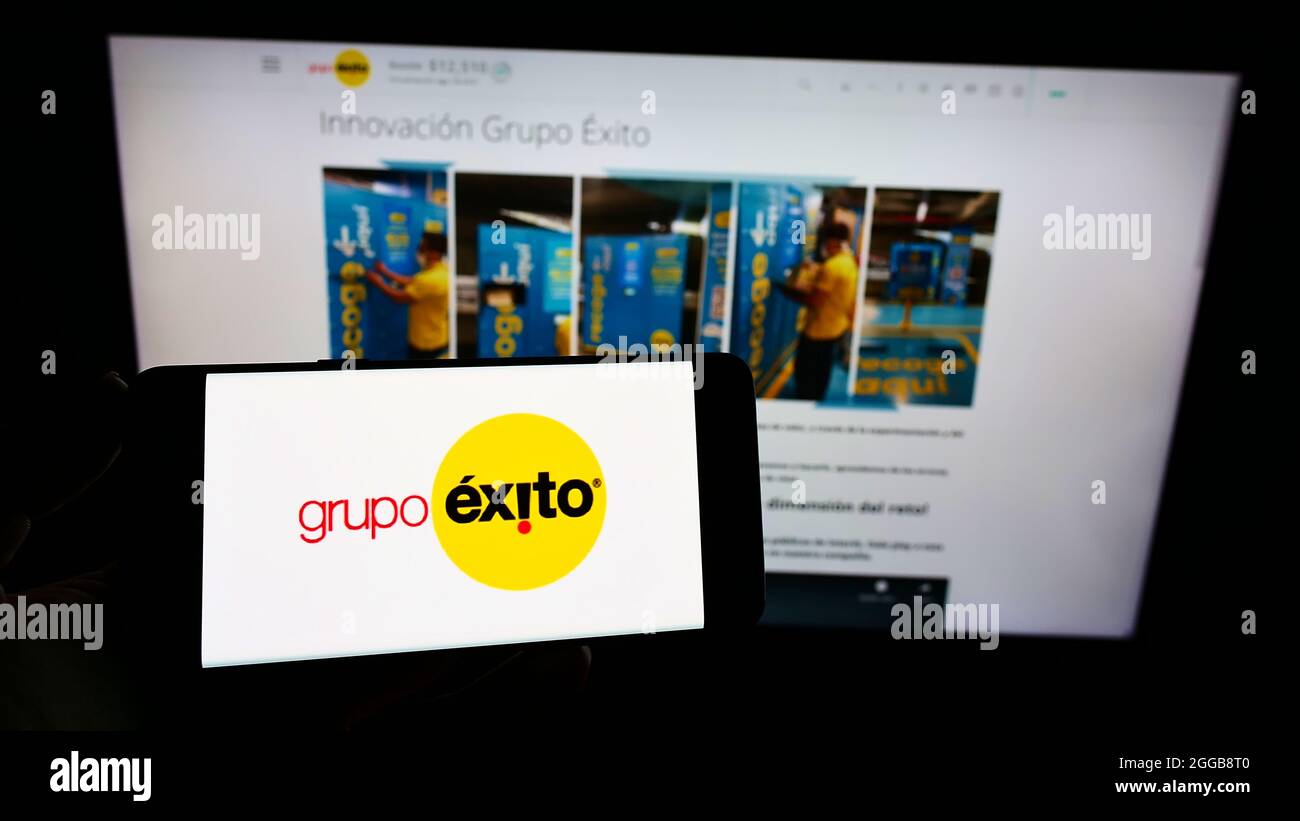 Person holding mobile phone with logo of company Almacenes Exito S.A. (Grupo Exito) on screen in front of web page. Focus on phone display. Stock Photo