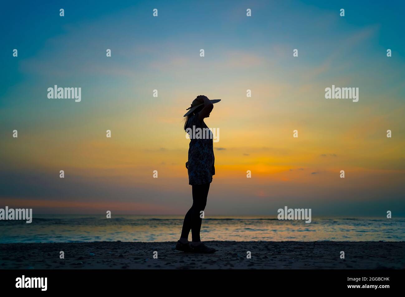 Side view silhouette of a young woman looking out to sea at sunset on a UK beach, staycation summer holiday, arms up holding on to her sun hat. Stock Photo