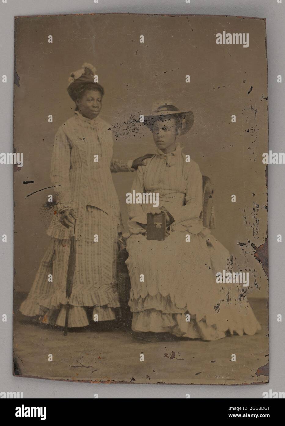 Untitled (Portrait of One Standing Woman and One Seated Woman), 1860s. Black-and-white  portrait photograph of two women with medium-dark skin, one sitting and the  other standing. They both wear light-colored, striped, ruffled
