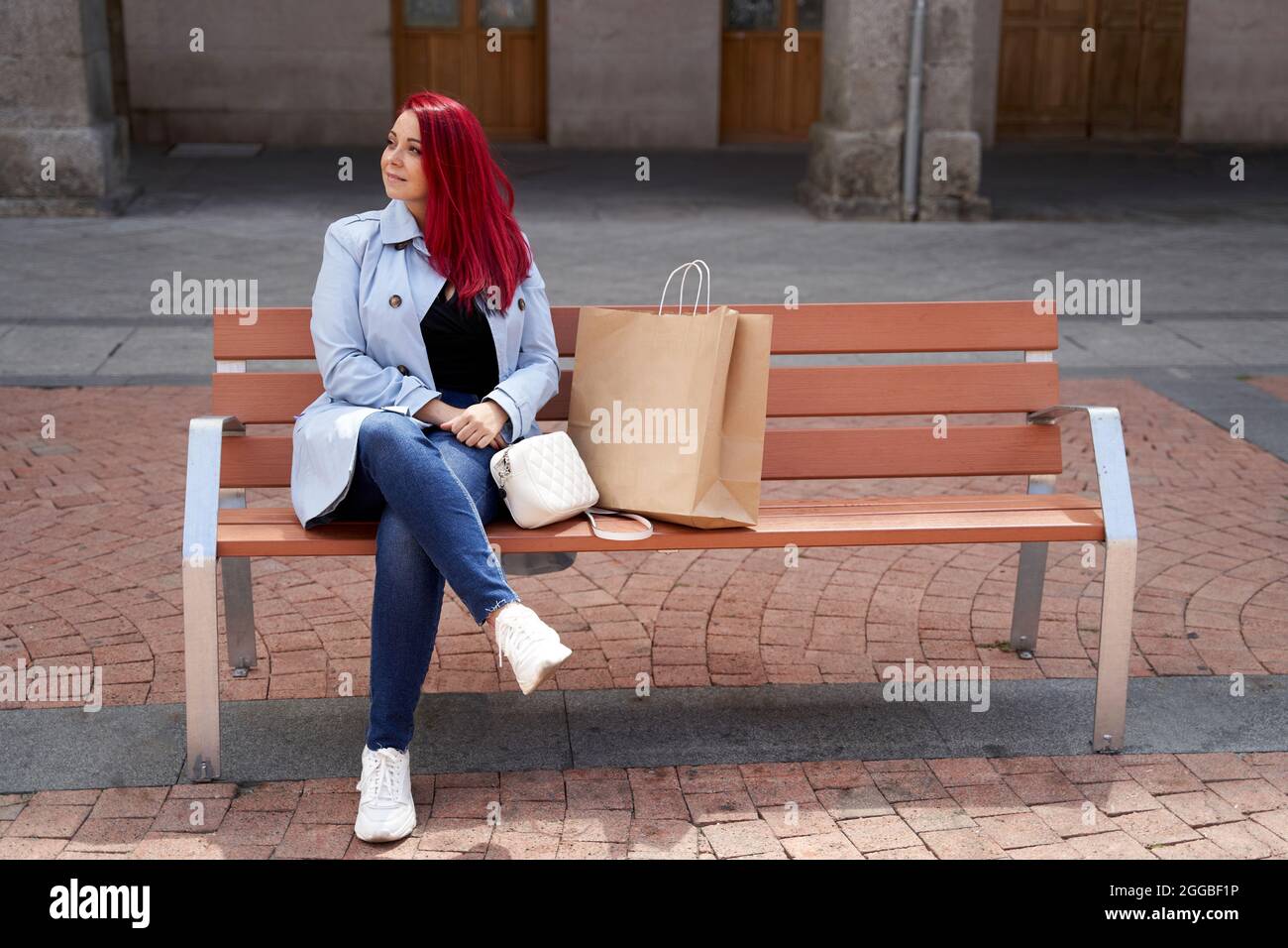 Red-haired woman in a blue trench coat sitting on a bench in the square ...