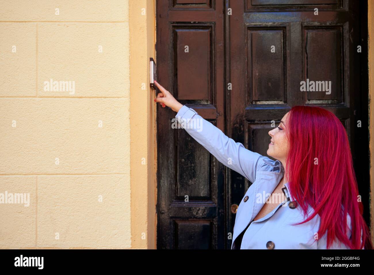 Red-haired woman in blue trench coat calling the intercom of a house. High quality photo Stock Photo