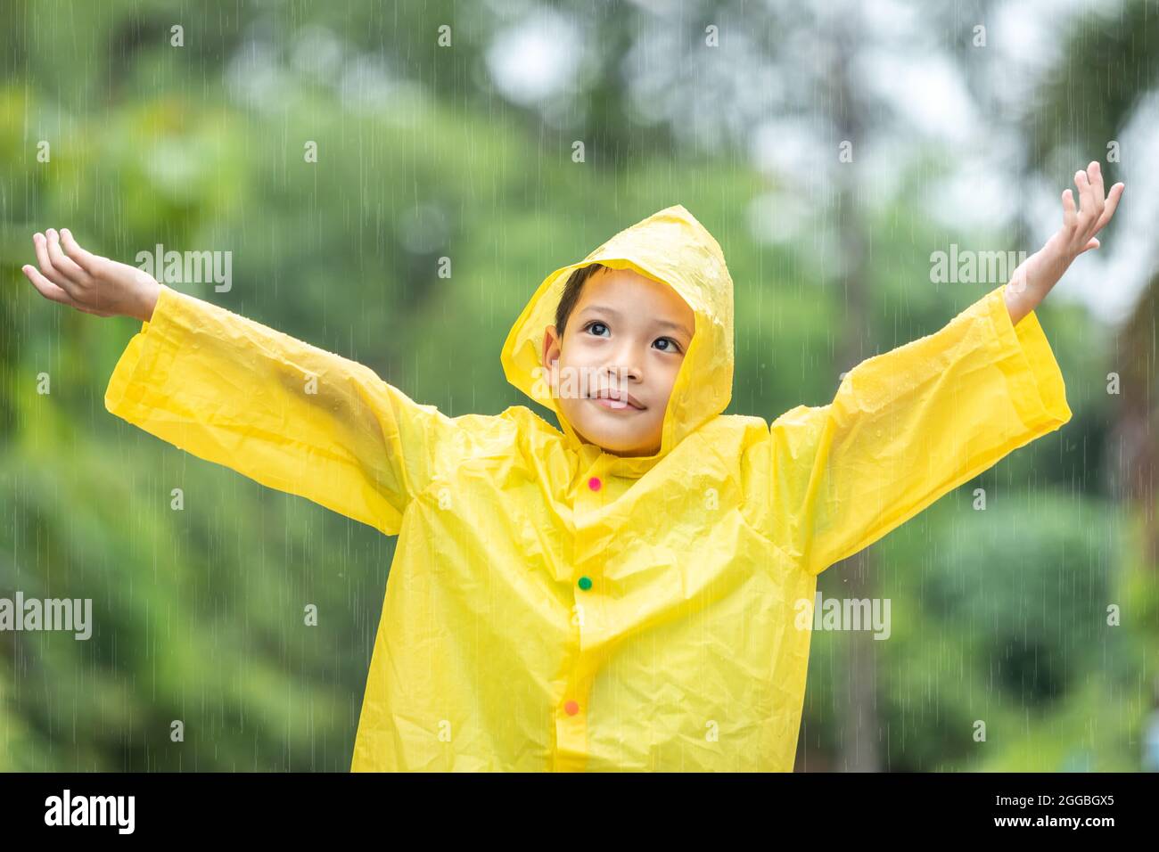 A boy wearing a yellow raincoat. Happy Asian little child having fun playing with the raindrops. A boy looking up at the sky and enjoying rainfall. Stock Photo
