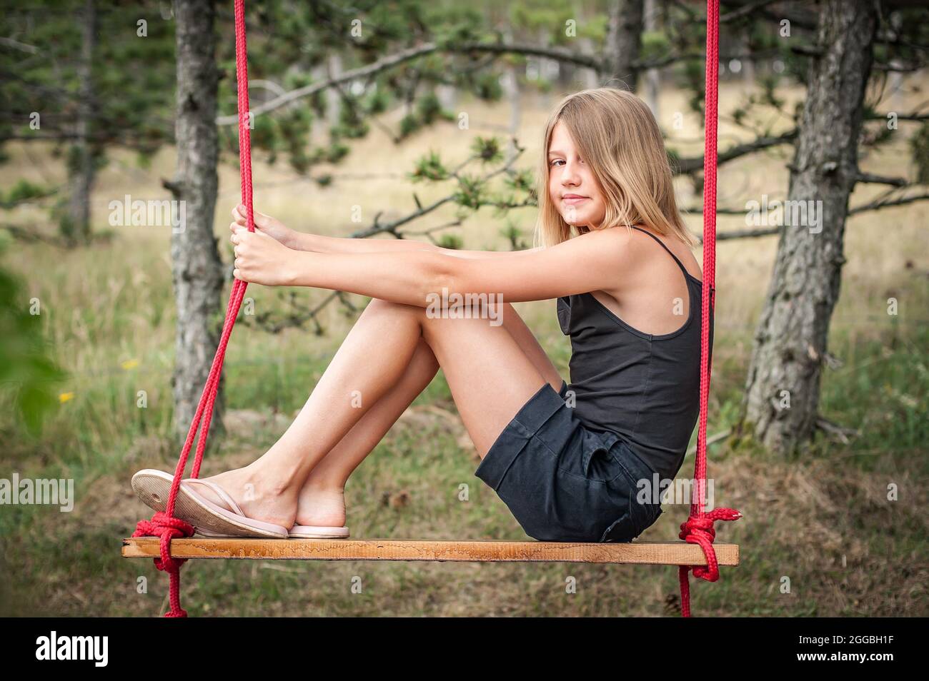 Beautiful teenage girl sitting and enjoying on swing in summer pine forest. Childhood, lifestyle concept Stock Photo