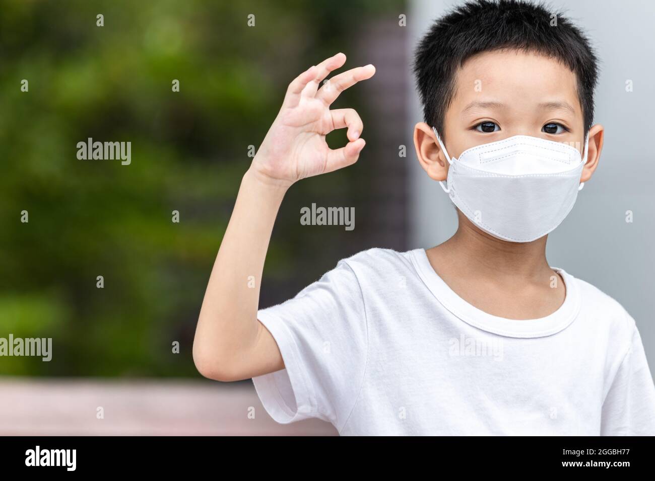 Cute smiling little child boy making OK gesture. Happy Asian boy in white t-shirt showing ok hand sign. A kid wearing face mask protection virus conce Stock Photo