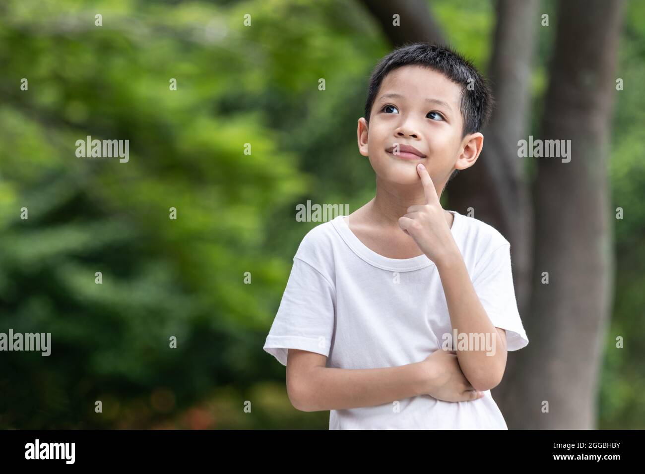 A child wearing a white t-shirt looks smart-looking up in doubt. Little Asian boy thinking with hand on chin. Stock Photo