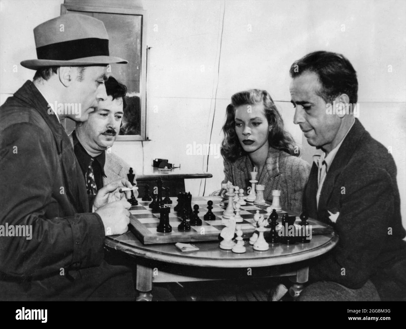 CHARLES BOYER plays Chess with Set Visitor HUMPHREY BOGART while former Chess Champion and Set Visitor HERMAN STEINER and LAUREN BACALL watch in on set candid taken during filming of CONFIDENTIAL AGENT 1945 director HERMAN SHUMLIN novel Graham Greene screenplay Robert Buckner Warner Bros. Stock Photo