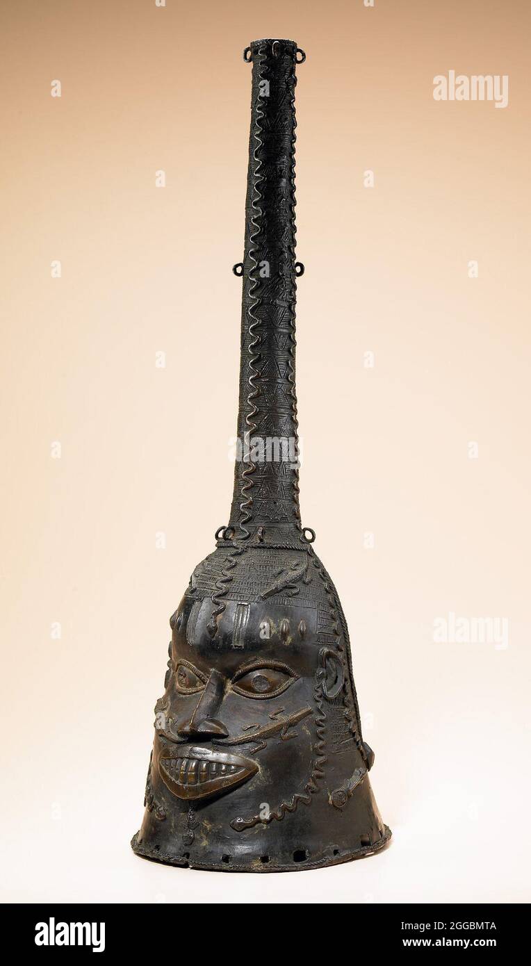 Cast copper alloy cone shaped mask with a human face, crocodiles issuing from the nostrils and snakes descending from the top projection. A relief of mud fish is scattered on the lower portion of the head and the mask is inlaid with iron in the eyes and in two vertical bars on the forehead. The rim of the neck opening is edged with a herringbone braid. Stock Photo