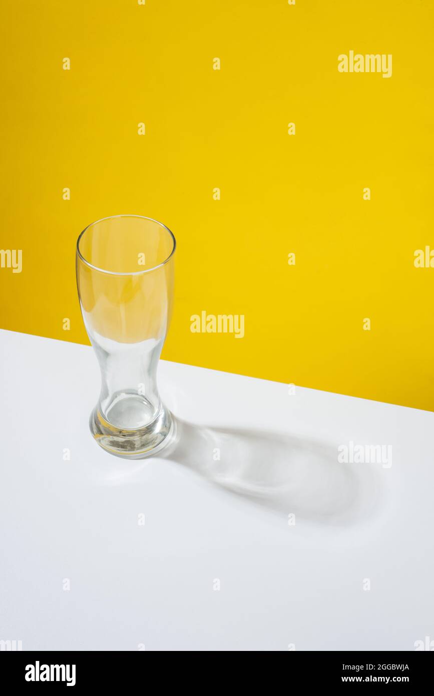 An empty beer glass placed on a white table in front of a yellow background Stock Photo