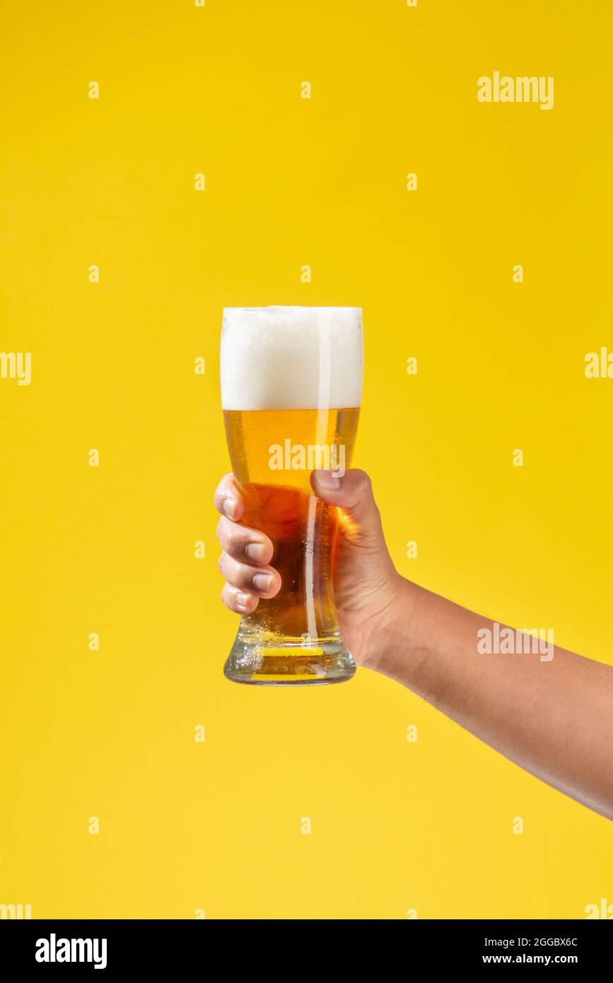 man's arm holds a glass beaker with beer inside, white foam and a solid yellow background Stock Photo