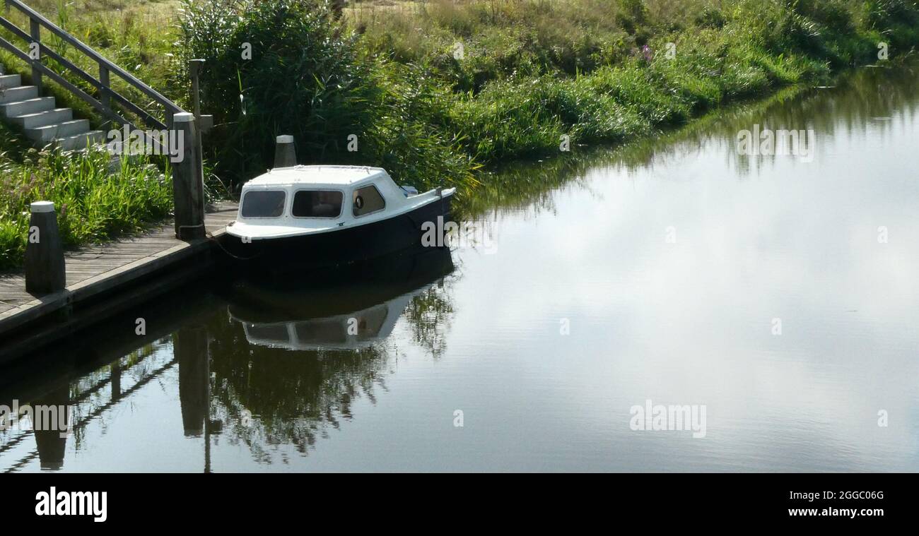 A small pleasure boat with cabin is anchored at a jetty. The water is a river in the Netherlands. A concrete staircase leads to the vessel. Stock Photo