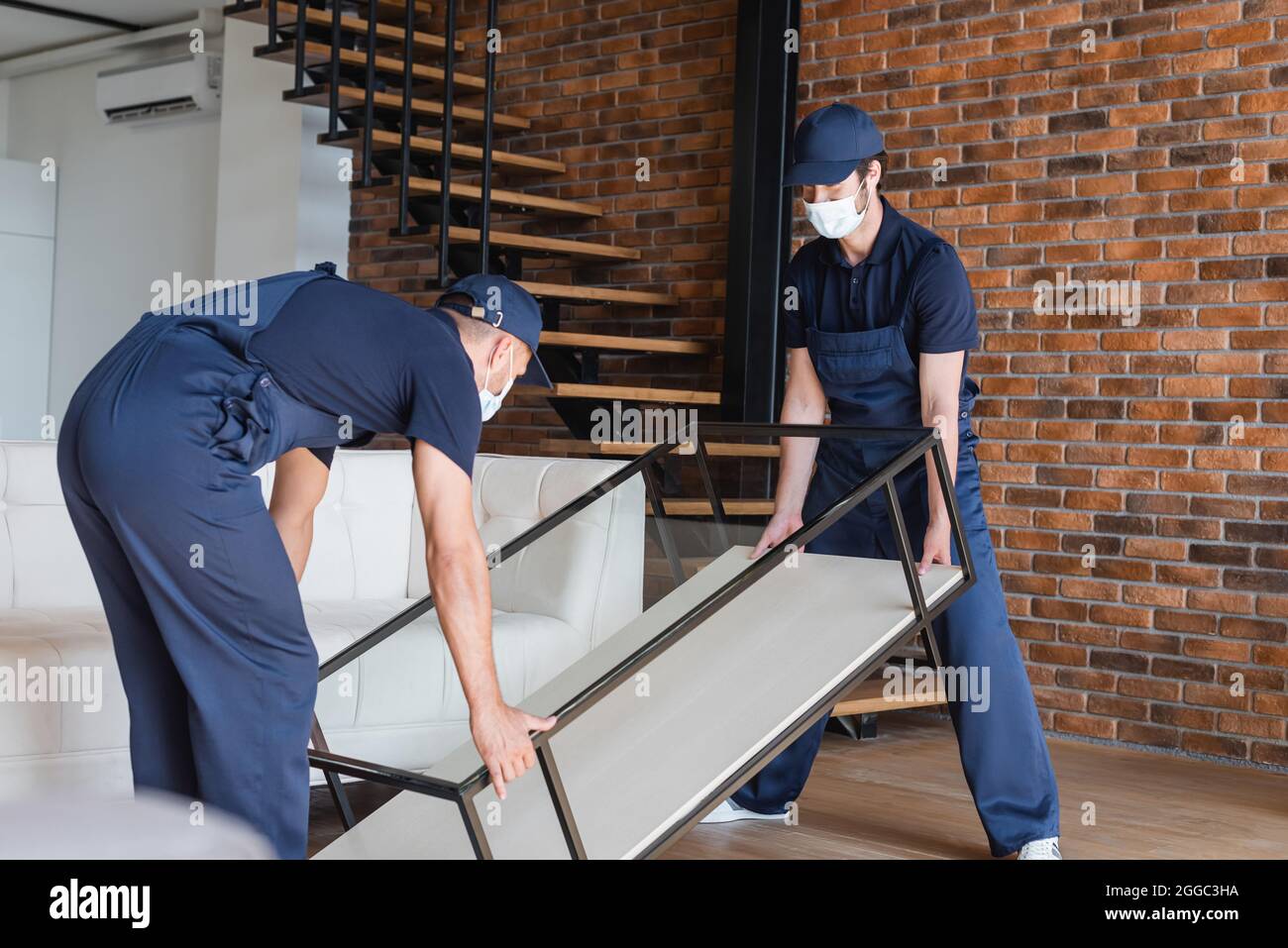 movers in uniform and medical masks lifting glass table near stairs
