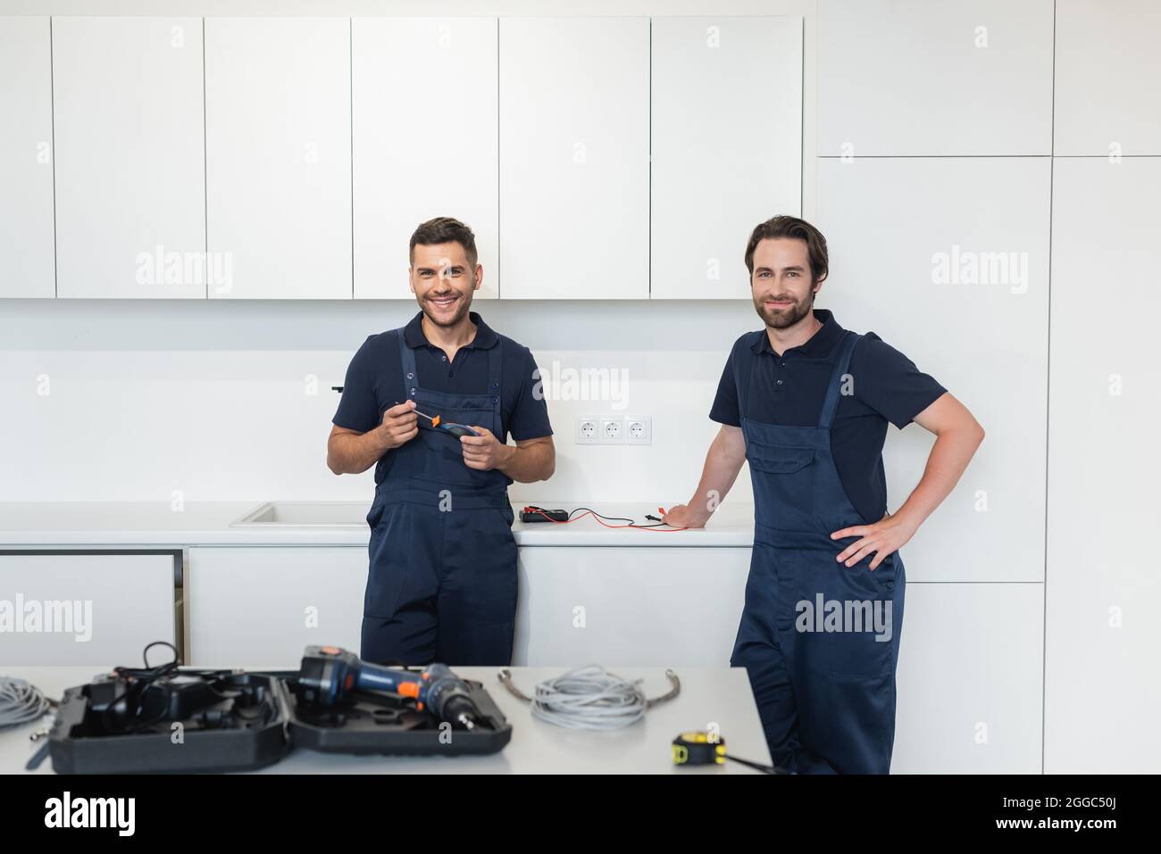 smiling handymen looking at camera near tools on kitchen table Stock Photo