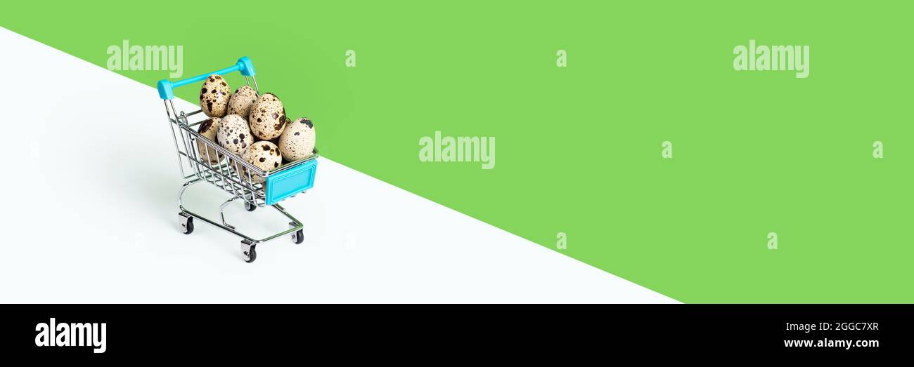 Toy supermarket cart or trolley filled with quail eggs isolated on green background. Long banner with copy space Stock Photo