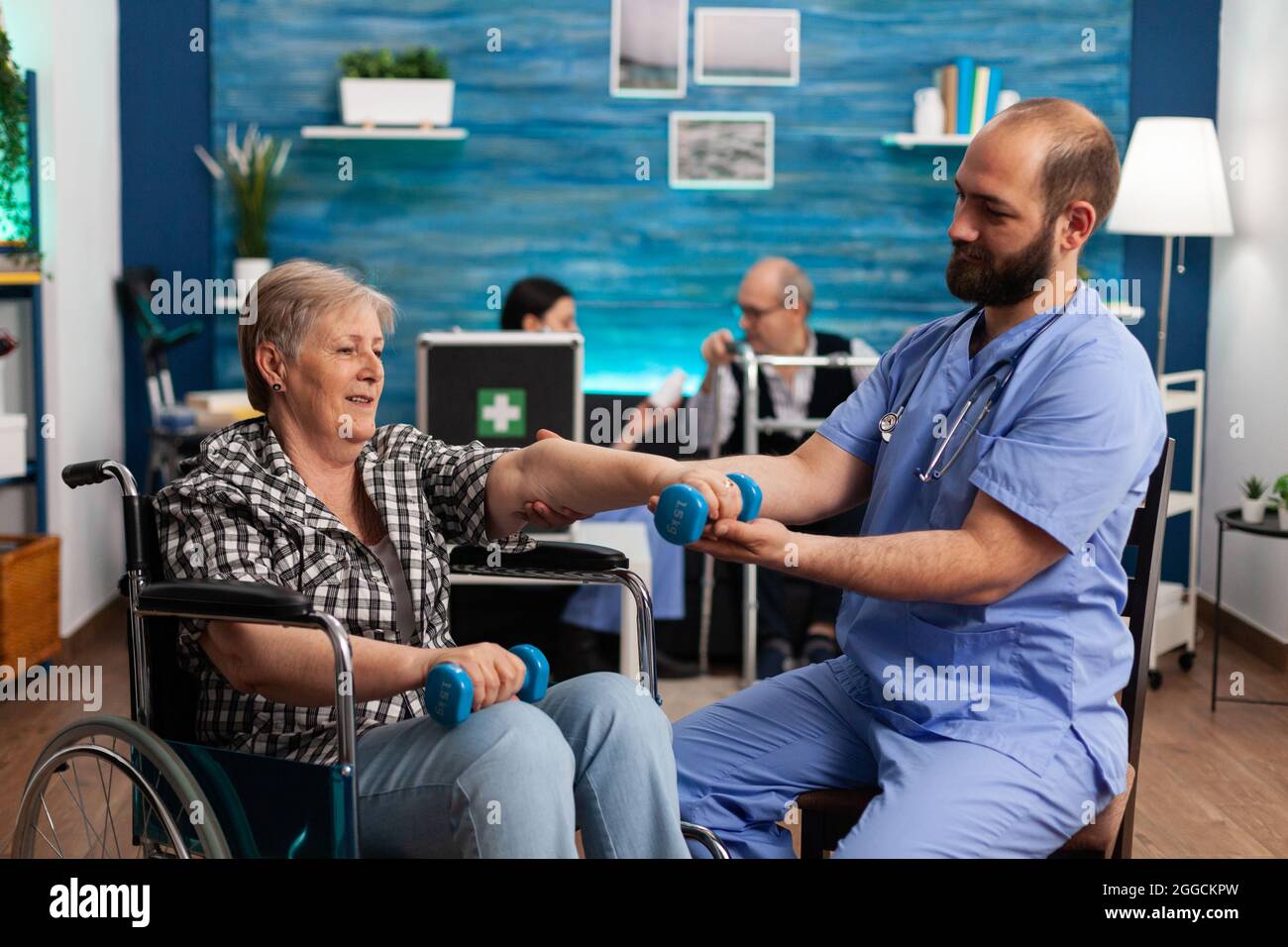 Male nurse helping senior retired disabled woman in wheelchair to rehabilitate using dumbbels during recovery session in nursing home. Elderly woman exercising physiotherapy with social worker Stock Photo