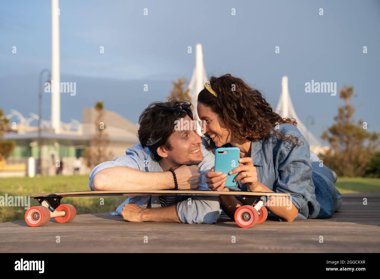 Carefree trendy couple surfing internet online outdoors use mobile phone internet lying on longboard Stock Photo