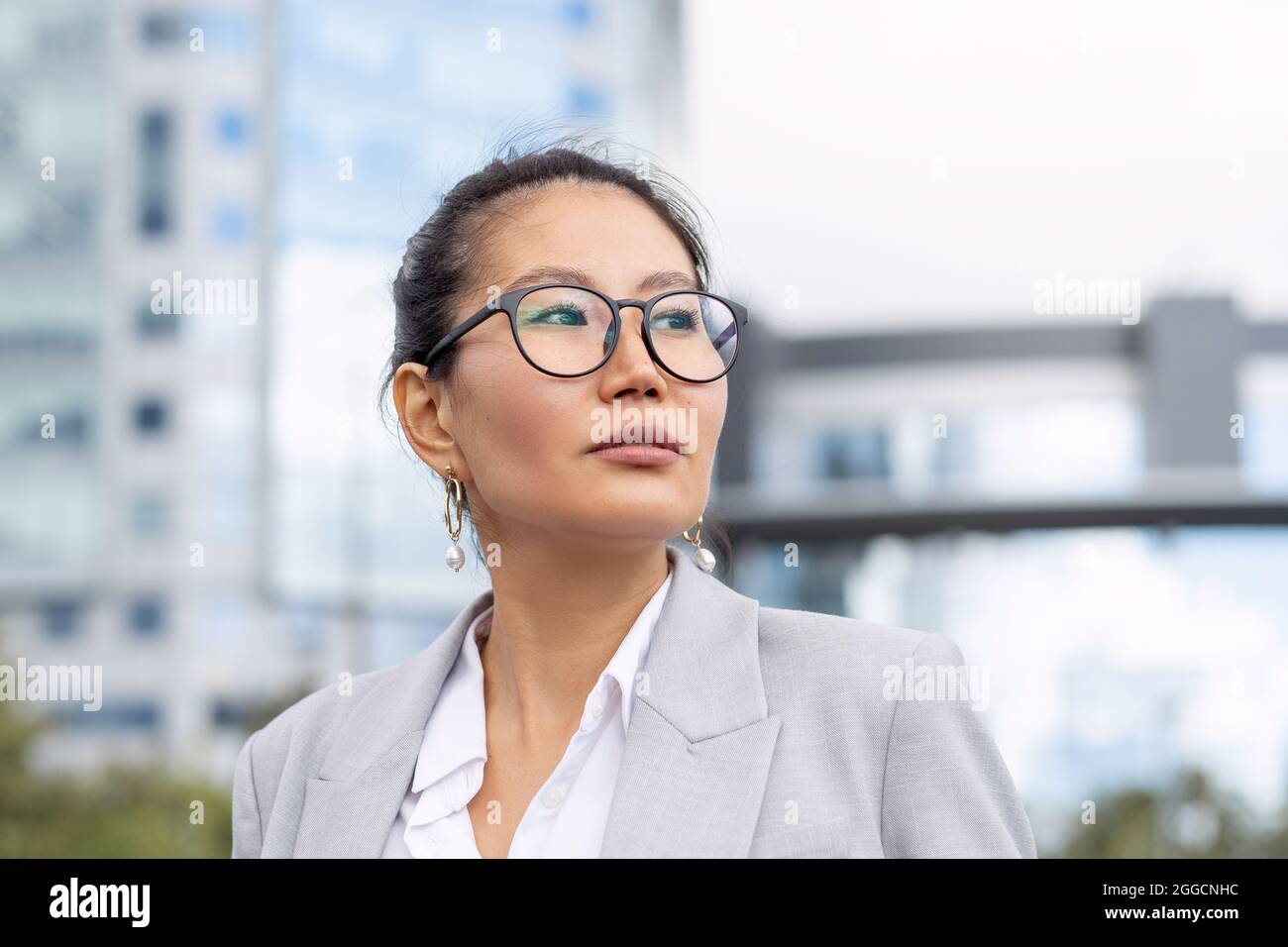 Young serious businesswoman of Asian ethnicity standing against modern architecture in urban environment Stock Photo