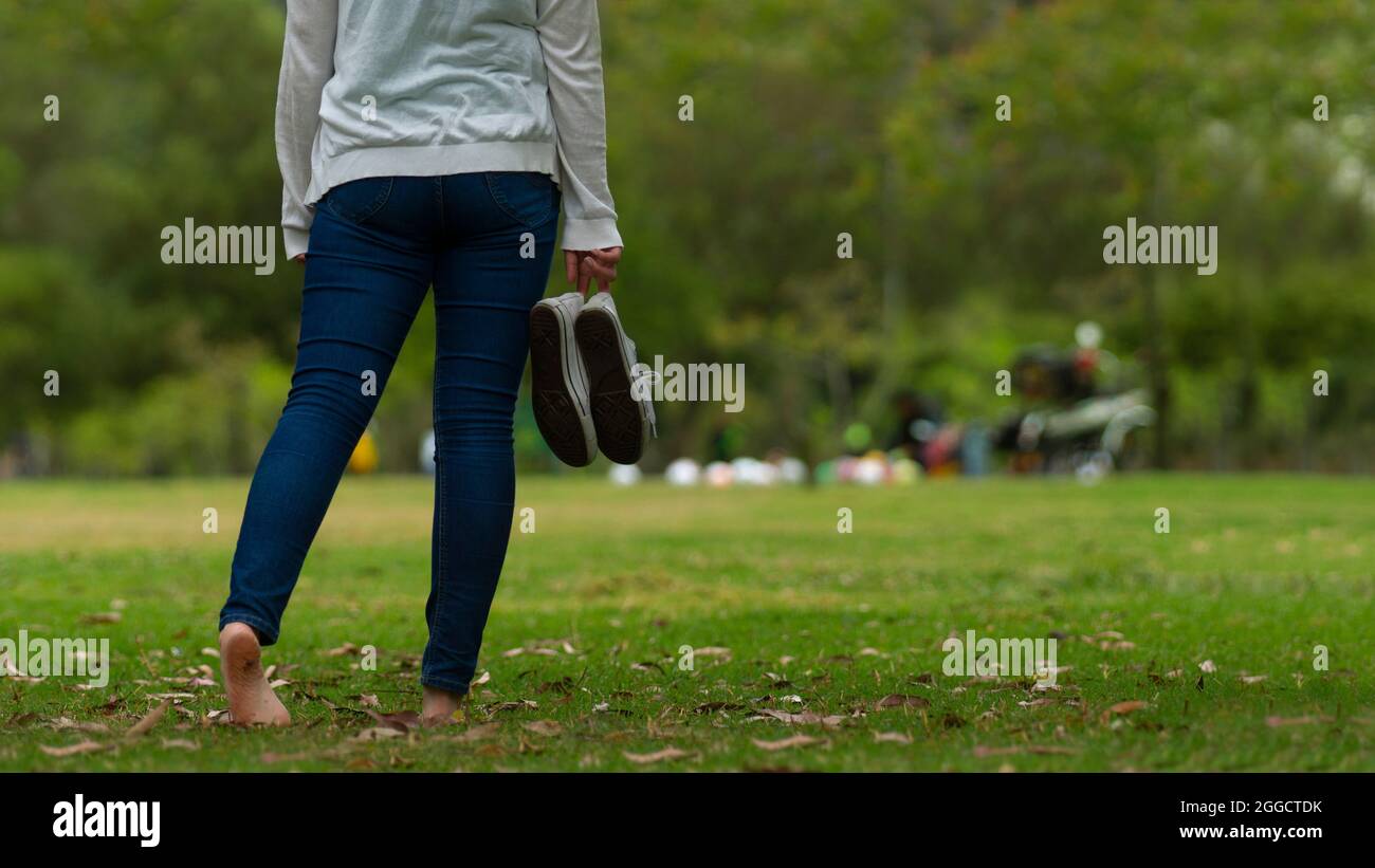 Closeup to young woman dressed in blue pants walking barefoot on the grass in the park, holding her sports shoes in her right hand with green trees ba Stock Photo