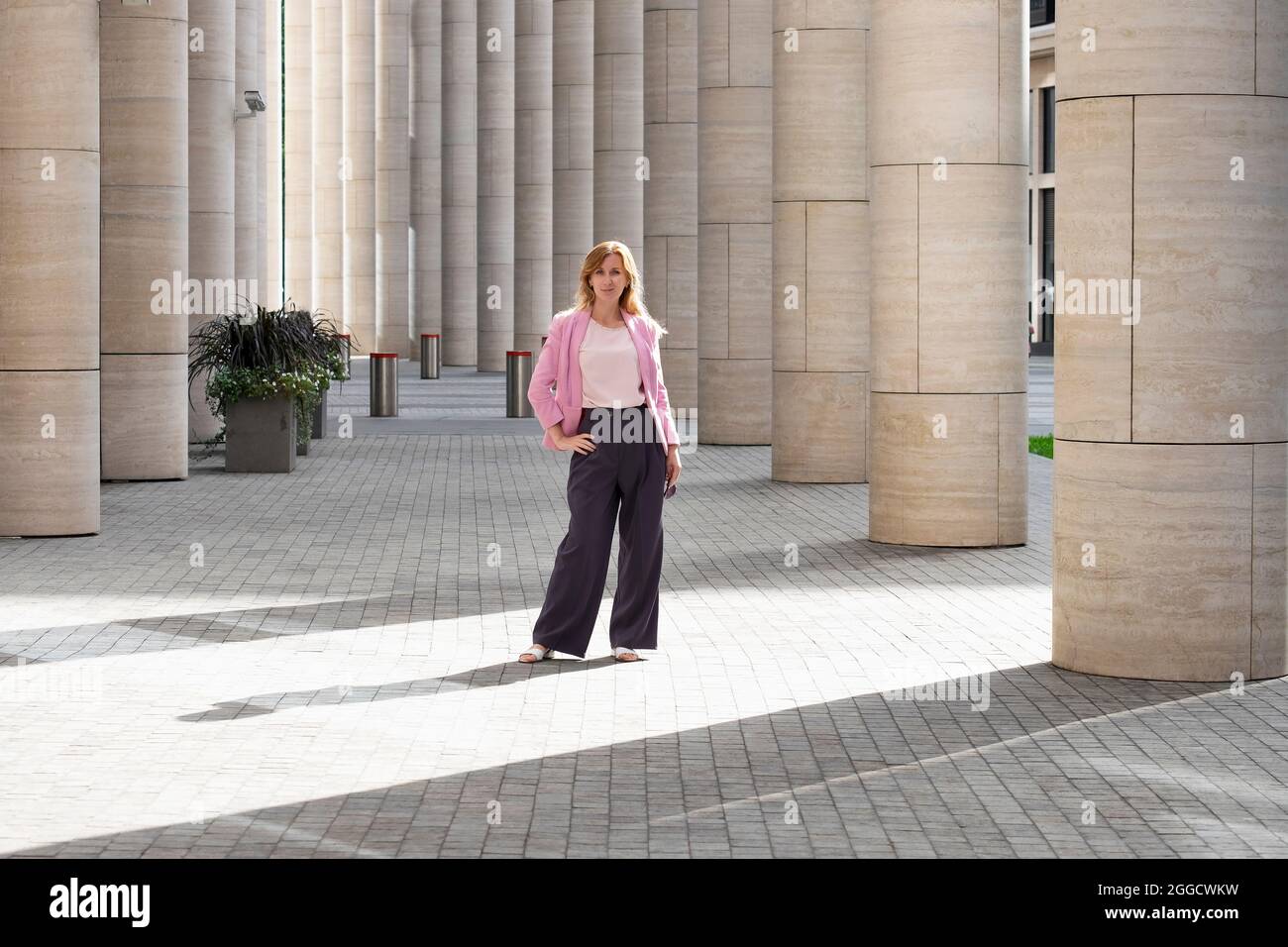 Young elegant beautiful blonde millennial with long hair in pink clothes stands in the colonnade of modern building. Full height. Selective focus. Stock Photo