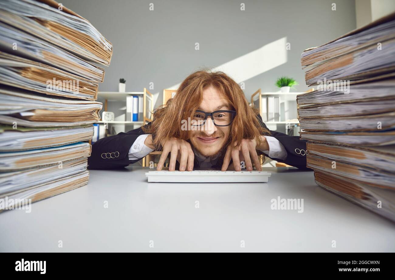 Happy office worker sitting at desk with stacks of papers and typing on wireless keyboard Stock Photo