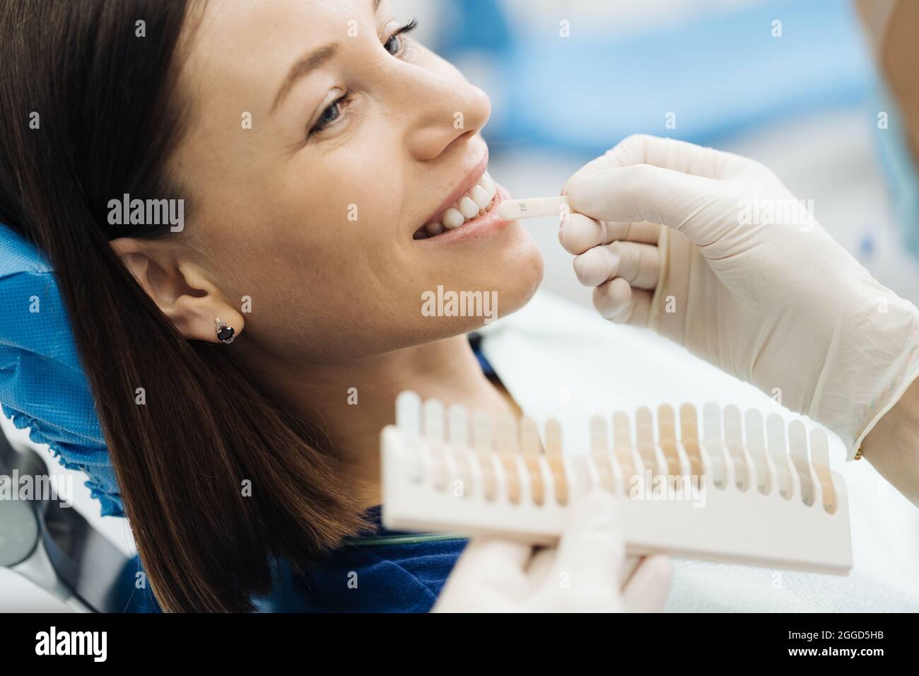 Close up portrait of beautiful young lady sitting in dental chair while stomatologist hands in sterile gloves holding tooth samples. She is smiling Stock Photo