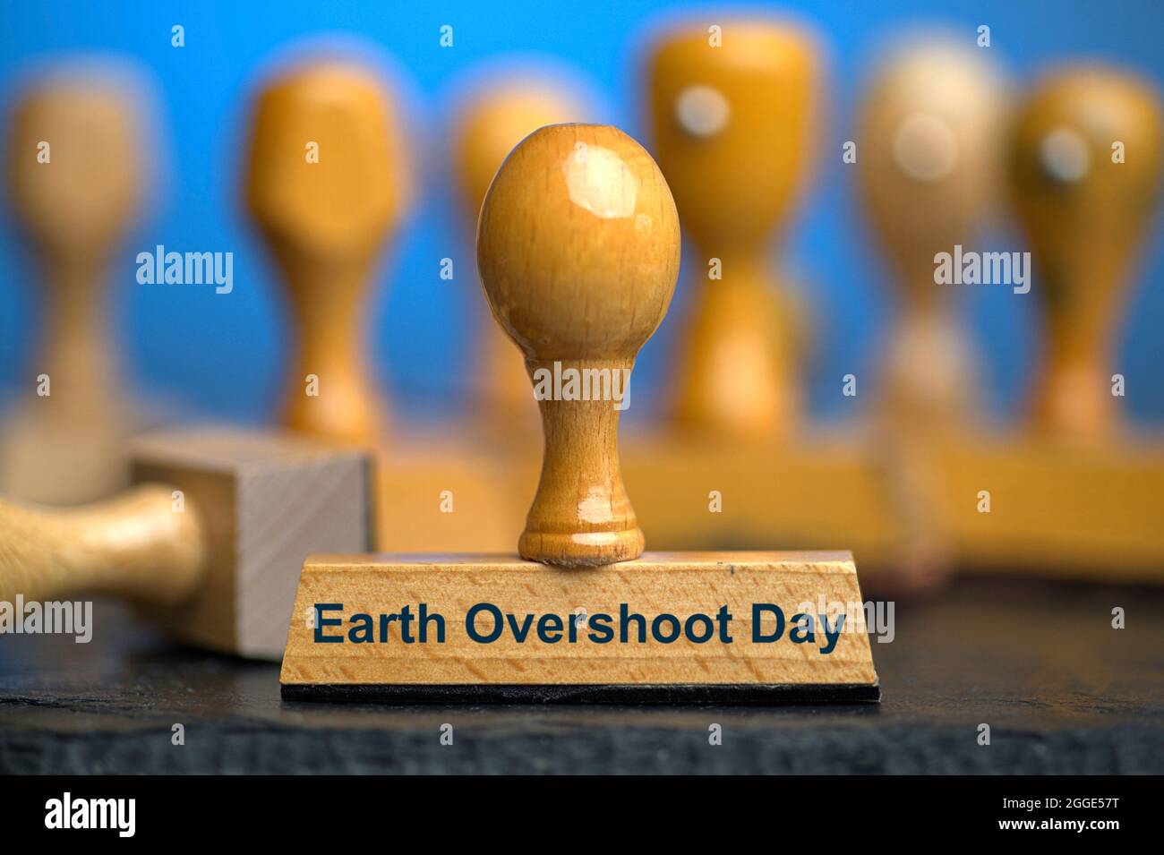 A wooden stamp with the inscription 'Earth Overshoot Day' on a slate plate with further stamps out of focus behind it. Photo with composing against a blue background. The Earth Overshoot Day, also Earth Overload Day, Ecological Debt Day, World Creation Day, World Overload Day or Eco Debt Day, is the day of the current year in an annual campaign by the organization 'Global Footprint Network' on which the human demand for after-growing raw materials the supply and capacity of Earth to reproduce these resources exceeds this year. In 2021, the day falls on July 29th. Stock Photo