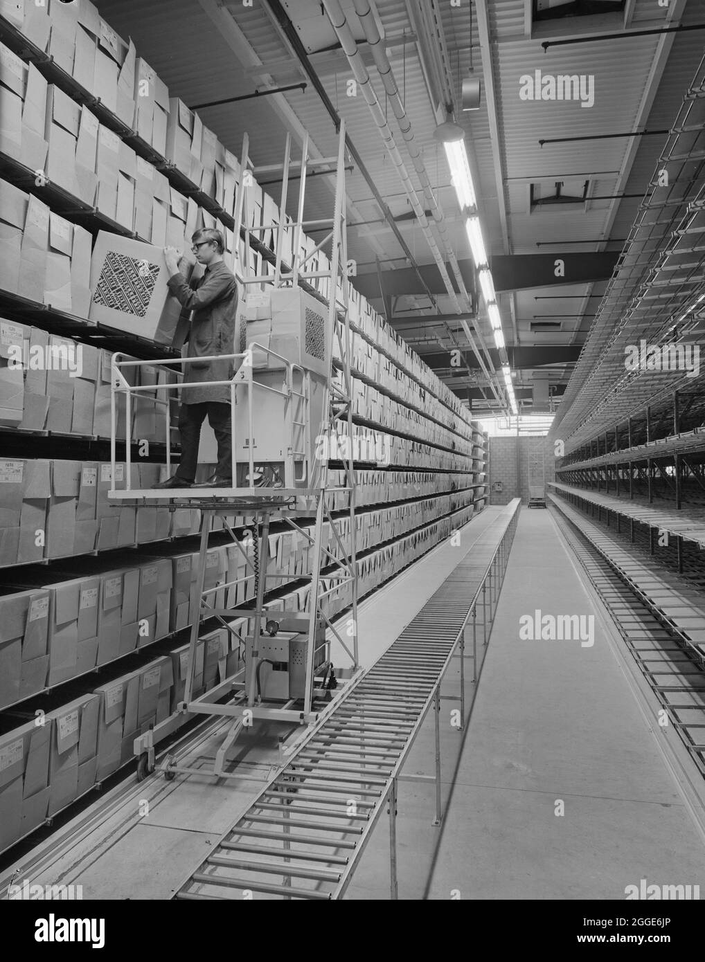 An interior view of the new Pirelli factory, showing shelving stacked with boxes and a man on a mobile platform lifting a box. The Pirelli factory in Carlisle was constructed by John Laing Construction Ltd. The factory was completed in two phases, the footwear division was first followed by the tyre division in November 1968. Stock Photo