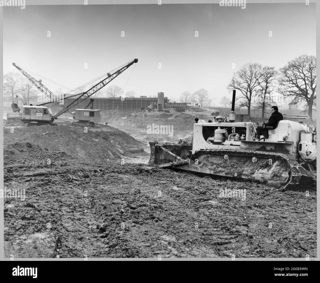 A view of the construction of the Stafford By-pass on the Birmingham to Preston Motorway (M6), looking north across Rising Brook towards the Highfields Farm access bridge and showing Laing excavators and a bulldozer carrying out peat removal. In the album, the typed caption below the photograph reads 'Looking north across Rising Brook where large pocket of peat (50,000 cu.yds) is being removed and replaced with clay backfill prior to the motorway embankment being constructed. Background shows Highfields Farm access bridge, which is virtually complete.' The work on the Stafford By-pass started Stock Photo