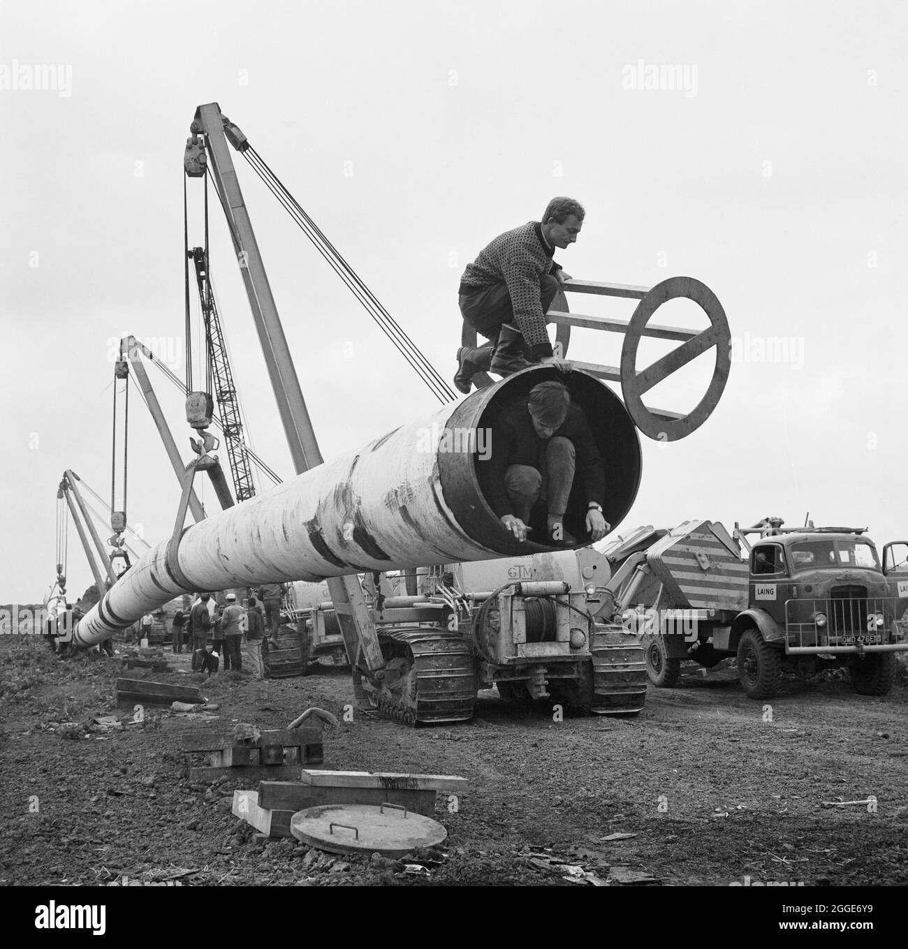 The Fens gas pipeline being lifted by a row of Caterpillar 583 pipelayers, with two workers crouched at one end of the pipe. Work on laying the Fens gas pipeline started in June 1967 and was a joint venture between Laing Civil Engineering and French companies Entrepose and Grands Travaux de Marseille (GTM) for the Gas Council. Over 600 men worked on the project to lay 36 inch diameter steel pipes starting at West Winch in Norfolk and running to where it linked up with the next contract at Woodcroft Castle in Cambridgeshire. The pipeline crossed four rivers and numerous dykes and ditches. This Stock Photo