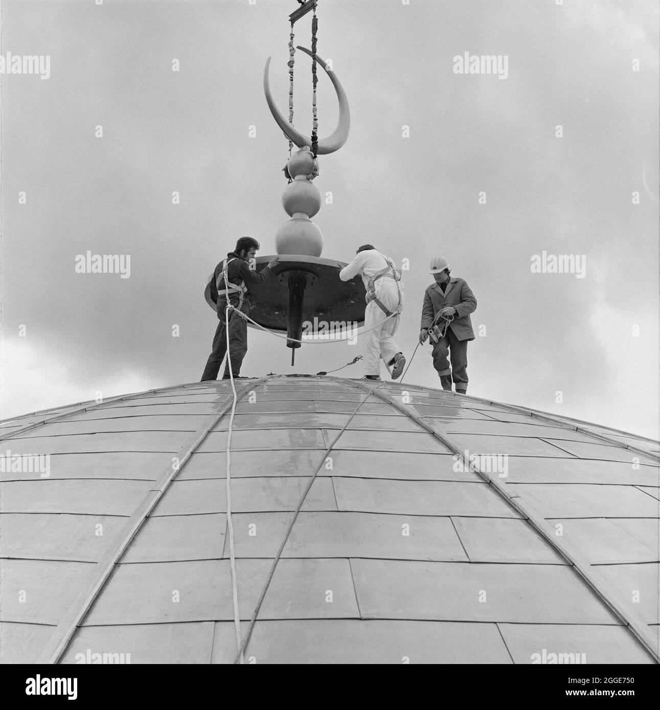 London Central Mosque and The Islamic Cultural Centre, Park Road, Regent's Park, City of Westminster, Greater London Authority, 15/03/1977. Men wearing safety harnesses positioning the crescent finial on the domed roof of the prayer hall at the London Central Mosque, as it is lowered from above by a crane. The finial was manufactured from glass-fibre reinforced concrete (GRC) in Northamptonshire, and painted in South Wales to give a gold coloured finish. Stock Photo