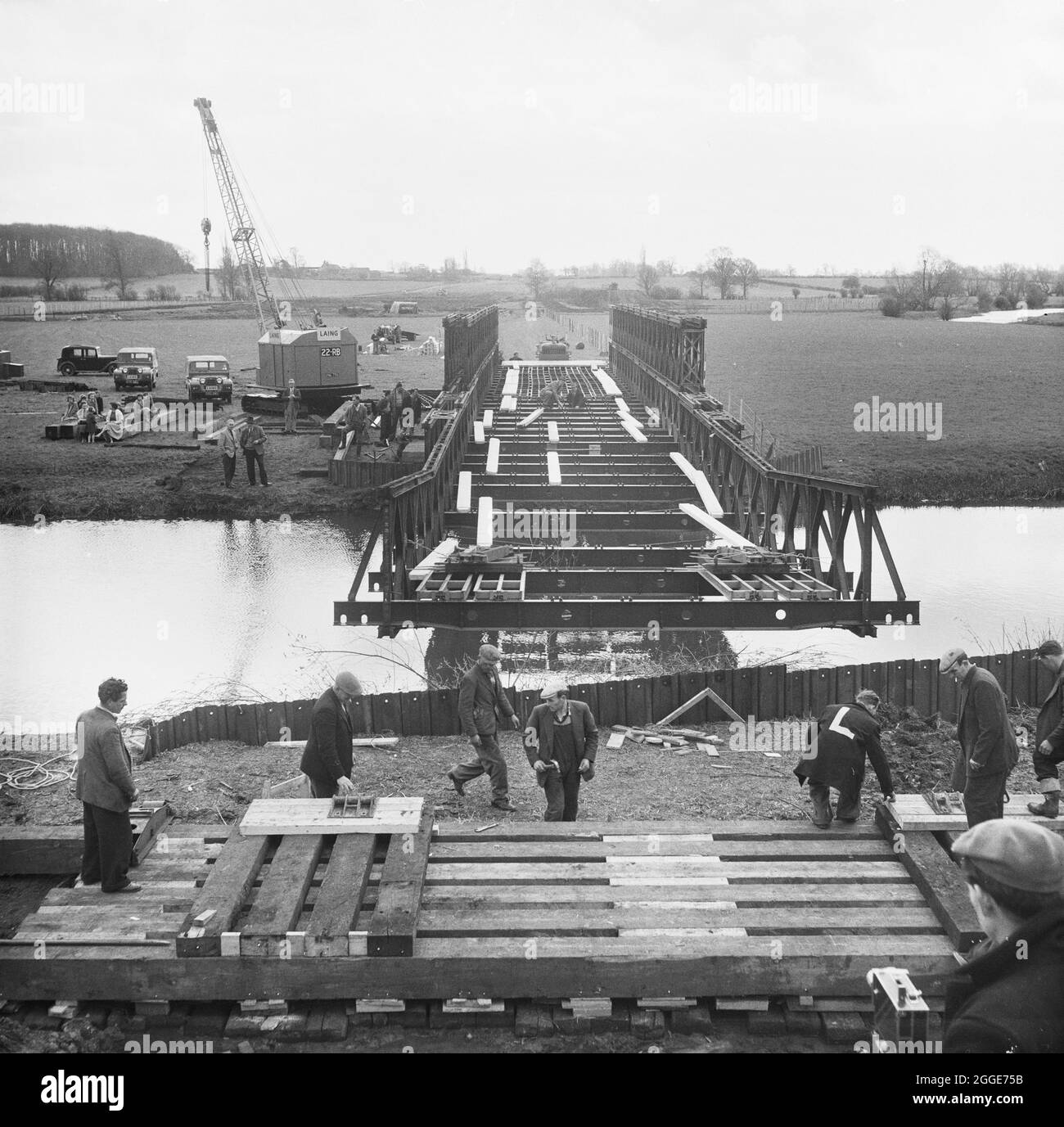 A Bailey bridge being erected across the main channel of the River Great Ouse near Newport Pagnell, during the construction of the London to Yorkshire Motorway (the M1). The caption beneath the corresponding album print states 'The Bailey Bridge being launched is a Double-double, effective span 100'0'', over the Main Ouse channel. 220,000 c/y's of excavated material will be transported over this bridge by Euclids, saving a six mile haul by road. In addition it also makes it possible to travel the full length of B3 Section on the line of the motorway'. Stock Photo