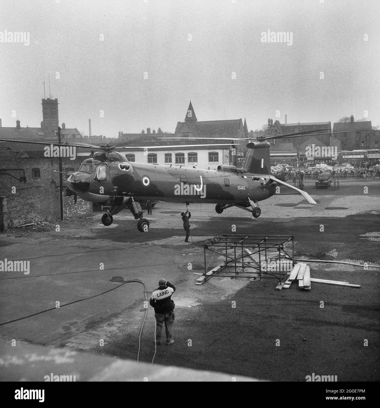 A RAF Belvedere helicopter hovering above tarmac just to the north of Coventry Cathedral, while a serviceman is attaching a hoist to the underside of the helicopter. A Laing worker is watching from the foreground and a crowd can be seen watching from the Pool Meadow Bus Station in the background. The photograph was taken during 'Operation Rich Man' a joint project involving Royal Air Force staff and Laing staff. This involved a RAF Belvedere helicopter hoisting in to place the 80ft bronze spire on to the new cathedral. The part of the operation to lower the 1/2 ton cross that sits on top of th Stock Photo