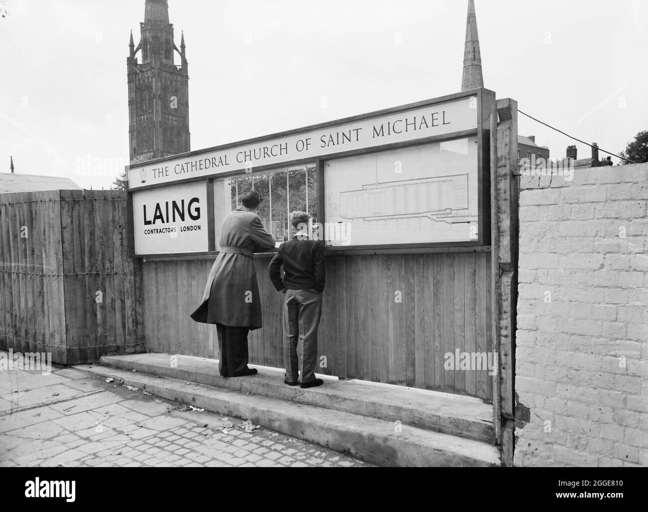 A man and boy looking through a viewing window at the construction site of Coventry Cathedral, with the Laing logo displayed on the left side, an elevation drawing of the cathedral displayed on the right side and a sign overhead displaying the cathedral name. This image was catalogued as part of the Breaking New Ground Project in partnership with the John Laing Charitable Trust in 2019-20. Stock Photo