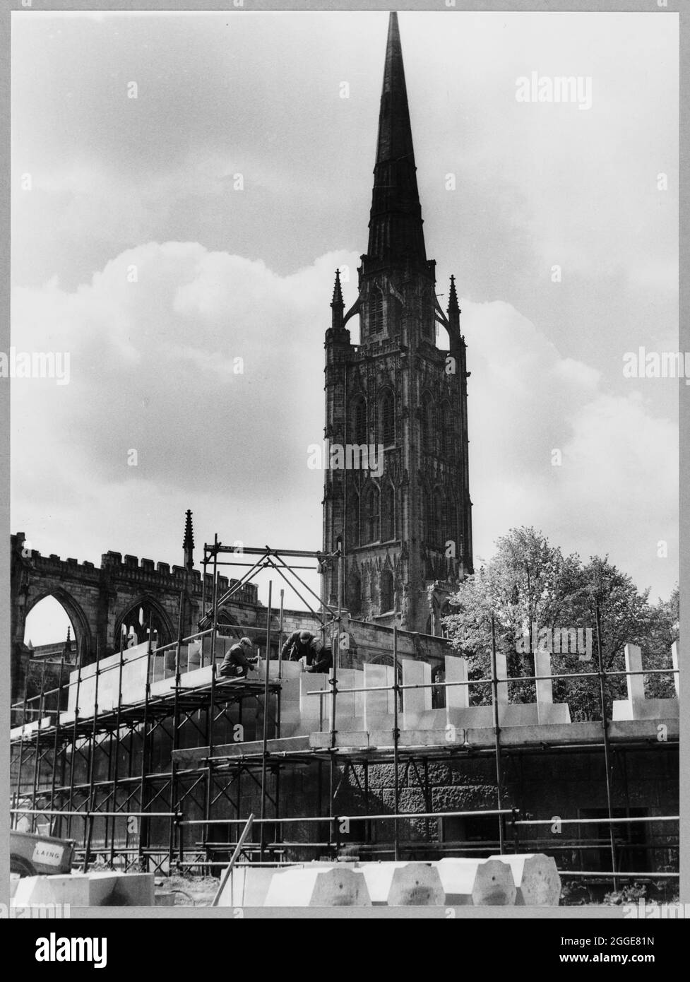 A view towards three builders working on a section of the baptistery window at Coventry Cathedral, showing some of the stones belonging to the window in the foreground and in the background the tower of the old cathedral. This image was catalogued as part of the Breaking New Ground Project in partnership with the John Laing Charitable Trust in 2019-20. Stock Photo