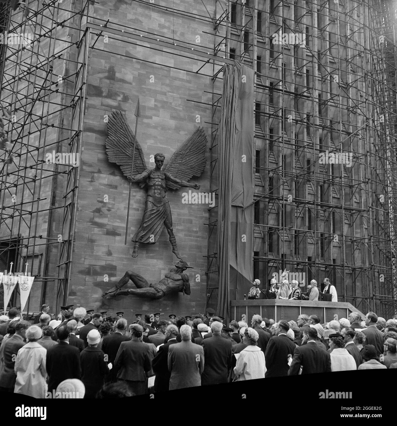 A crowd of people watching the unveiling ceremony of the statue St Michael and the Devil by sculptor Sir Jacob Epstein at Coventry Cathedral. The statue of the Archangel Michael overcoming the Devil was the last sculpture Sir Jacob Epstein created before he died in 1959. The statue overlooks the steps leading up to the entrance of Coventry Cathedral. The photograph shows the statue after it has been unveiled by his widow Lady Epstein, standing on the stage to the right. Stock Photo
