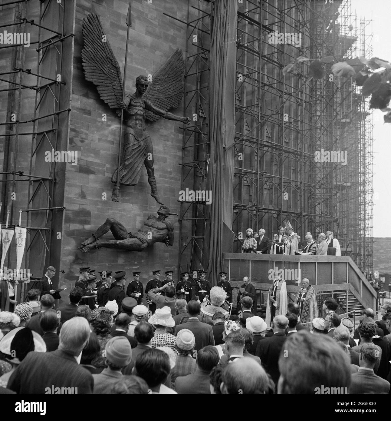 A crowd of people watching the unveiling ceremony of the statue St Michael and the Devil by sculptor Jacob Epstein at Coventry Cathedral. The statue of the Archangel Michael overcoming the Devil was the last sculpture Sir Jacob Epstein created before he died in 1959. The statue overlooks the steps leading up to the entrance of Coventry Cathedral. The photograph shows the statue after it has been unveiled by his widow Lady Epstein, standing on the stage to the right. Stock Photo