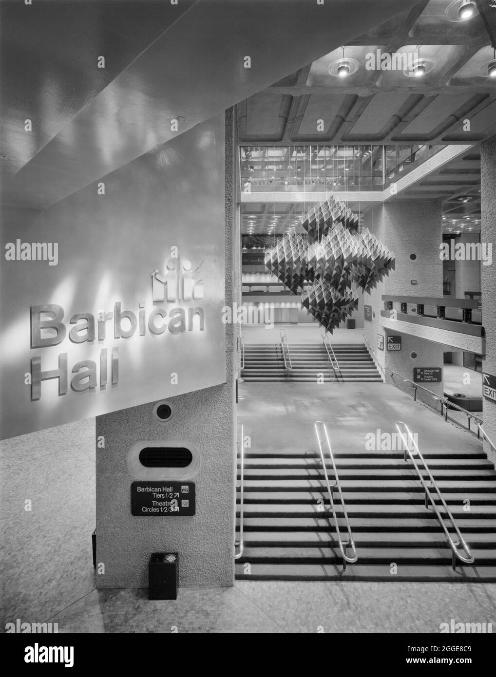 A view from a high vantage point of the main foyer in the newly opened Barbican Centre, showing a sculpture suspended over the staircase and the entrance for the Barbican Hall. This image was taken shortly after the opening of the Barbican Centre on Wednesday 3rd March 1982. The Barbican Centre was ceremonially opened by Queen Elizabeth II, who unveiled a commemorative plaque to mark the occasion. The sculpture in this image, designed by Michel Santry, is suspended from a skylight above the stairs. The sculpture is in the shape of an octahedron, and is built from six honeycomb groups of gold p Stock Photo