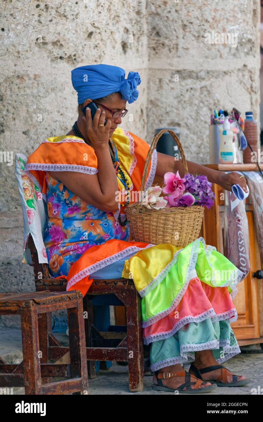 Woman, Cuban, in multicoloured traditional traditional costume, sitting on chair and talking on mobile phone, Old Town, Capital Havana, Havana Stock Photo