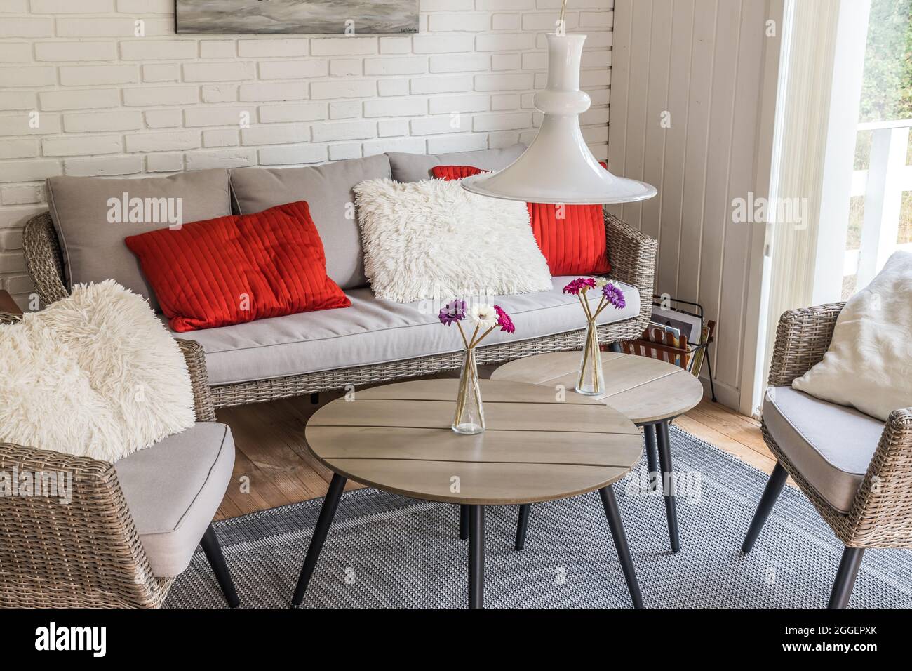 Rattan sofa with two armchairs and cozy red and white pillows, white stone wall behind, scandinavian style. Stock Photo