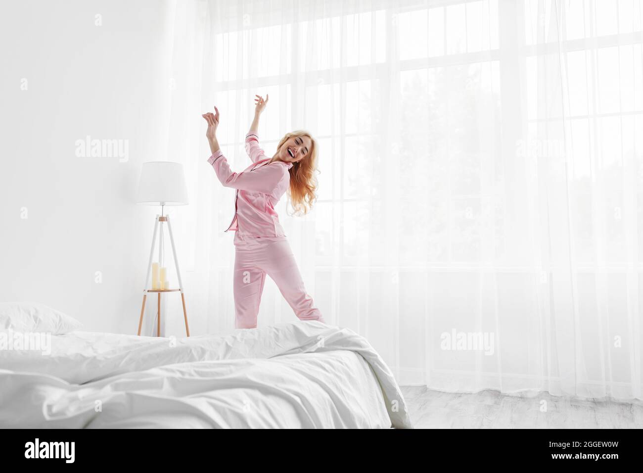 Beautiful blonde in pajama waking up in morning, happy young lady greets new day with good mood. Smiling beautiful european woman dancing near window Stock Photo