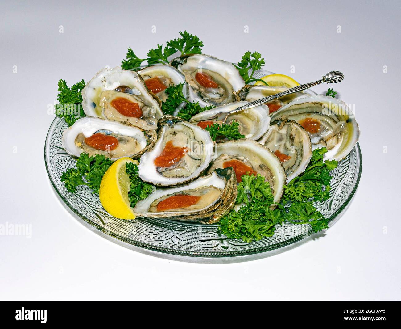 Round glass plate of freshly shucked oysters served with red cocktail sauce, yellow lemon and green curly parsley garnish. Stock Photo