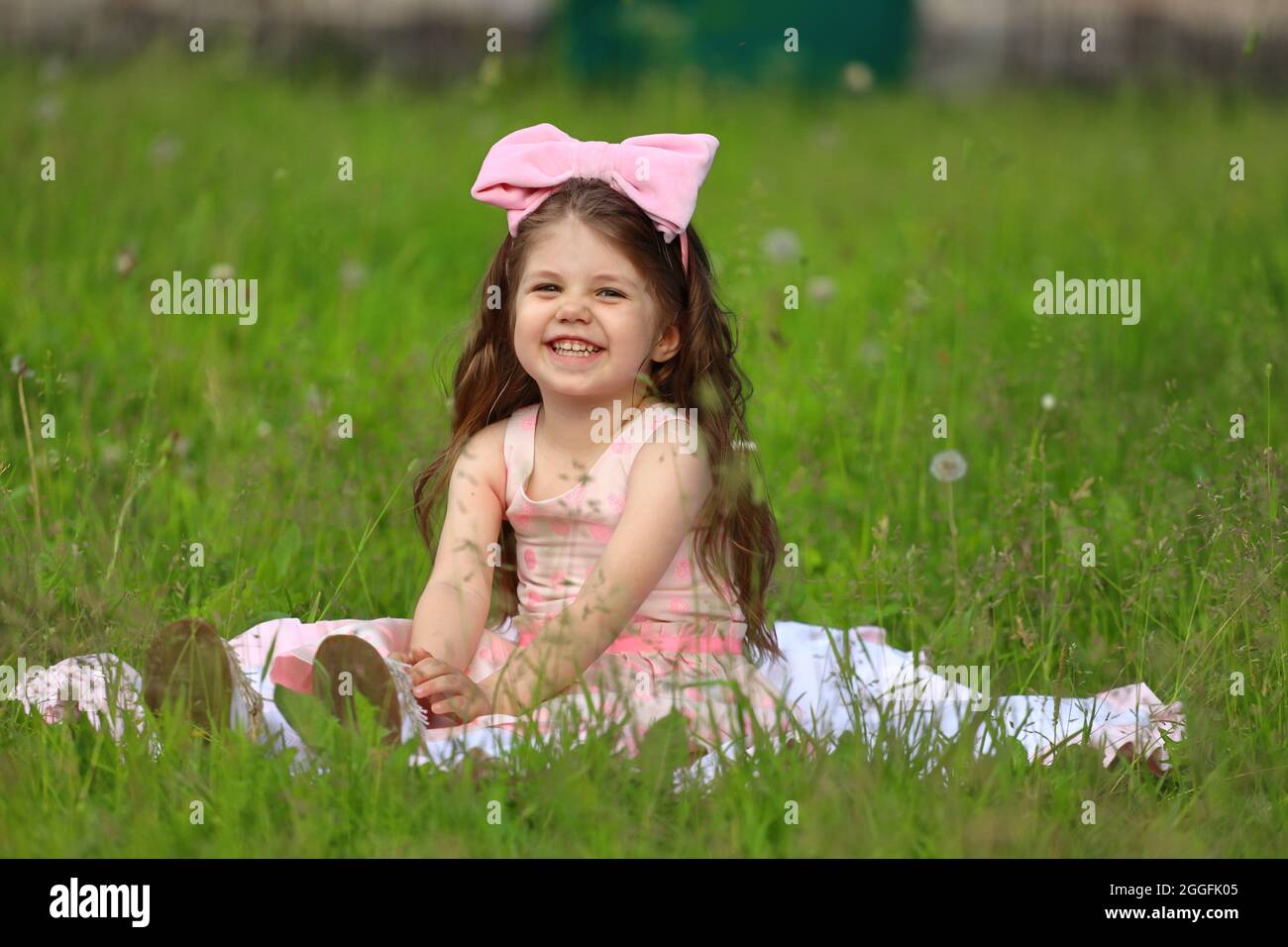 cute girl in a pink dress with a hat sits on the grass in the park ...