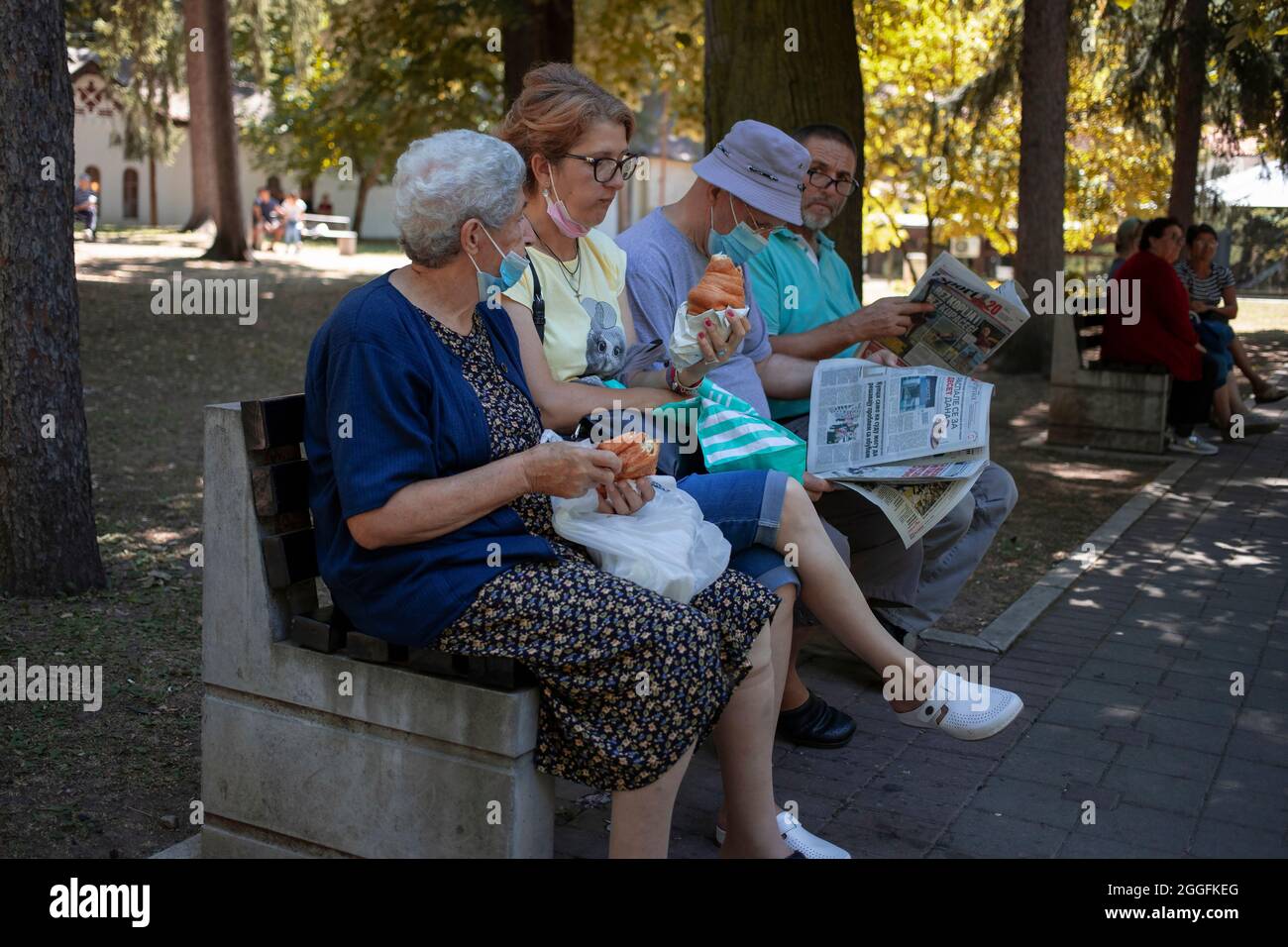 Sokobanja, Serbia, Aug 19, 2021: Two women seated on a bench having snack next to men reading newspaper Stock Photo