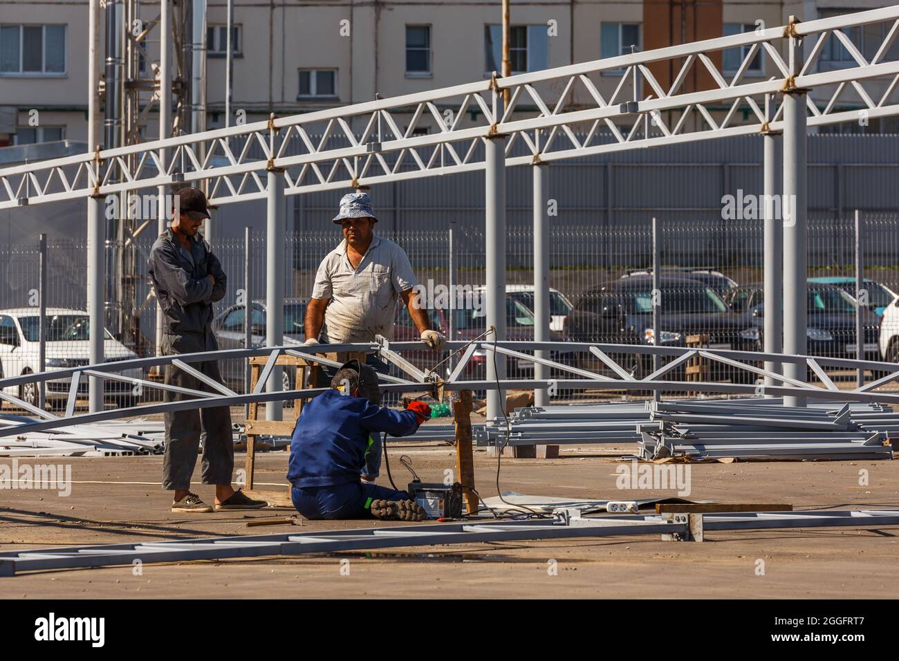 three Asian workers assembling metal construction by welding in front of car parking at sunny summer day under direct hard light - Tula, Russia, 08 30 Stock Photo