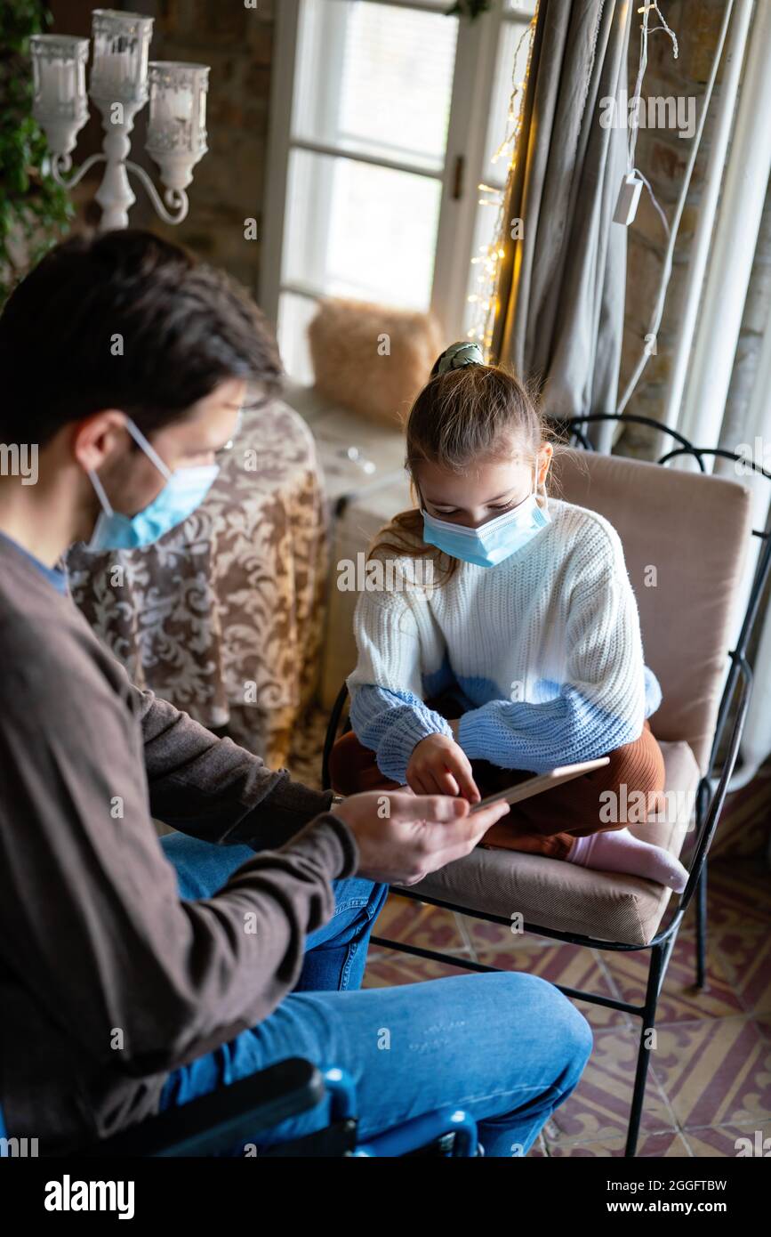 Father with disability in wheelchair using tablet at home with child while wearing masks. Stock Photo