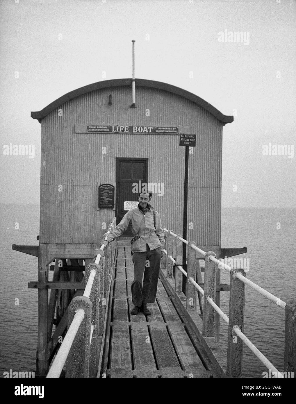 1950s, historical, a man standing on the wooden gangway in front of the old lifeboat station at Selsey, West Sussex, England, UK. A lifeboat service was established in Selsey in 1861. In 1927 to house the station's new motor lifeboat, the boat house was re-built and this structure is seen in the picture. This slipway station was again rebuilt in the late 1950s but was taken down in 2017. Stock Photo