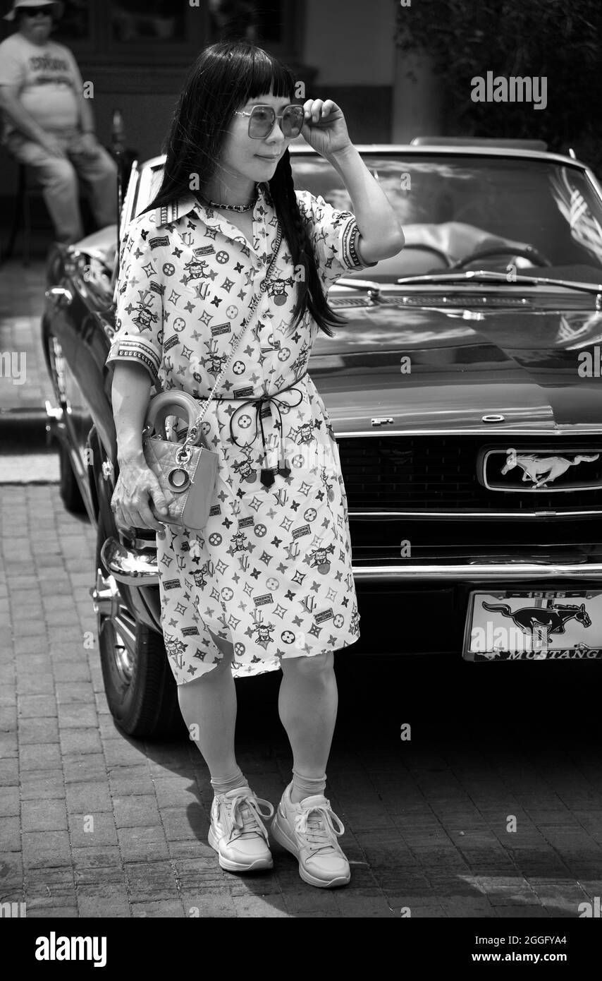 A Chinese woman wearing a Louis Vuitton dress and carrying a Christian Dior handbag stands beside a vintage Ford Mustang in Santa Fe, New Mexico. Stock Photo