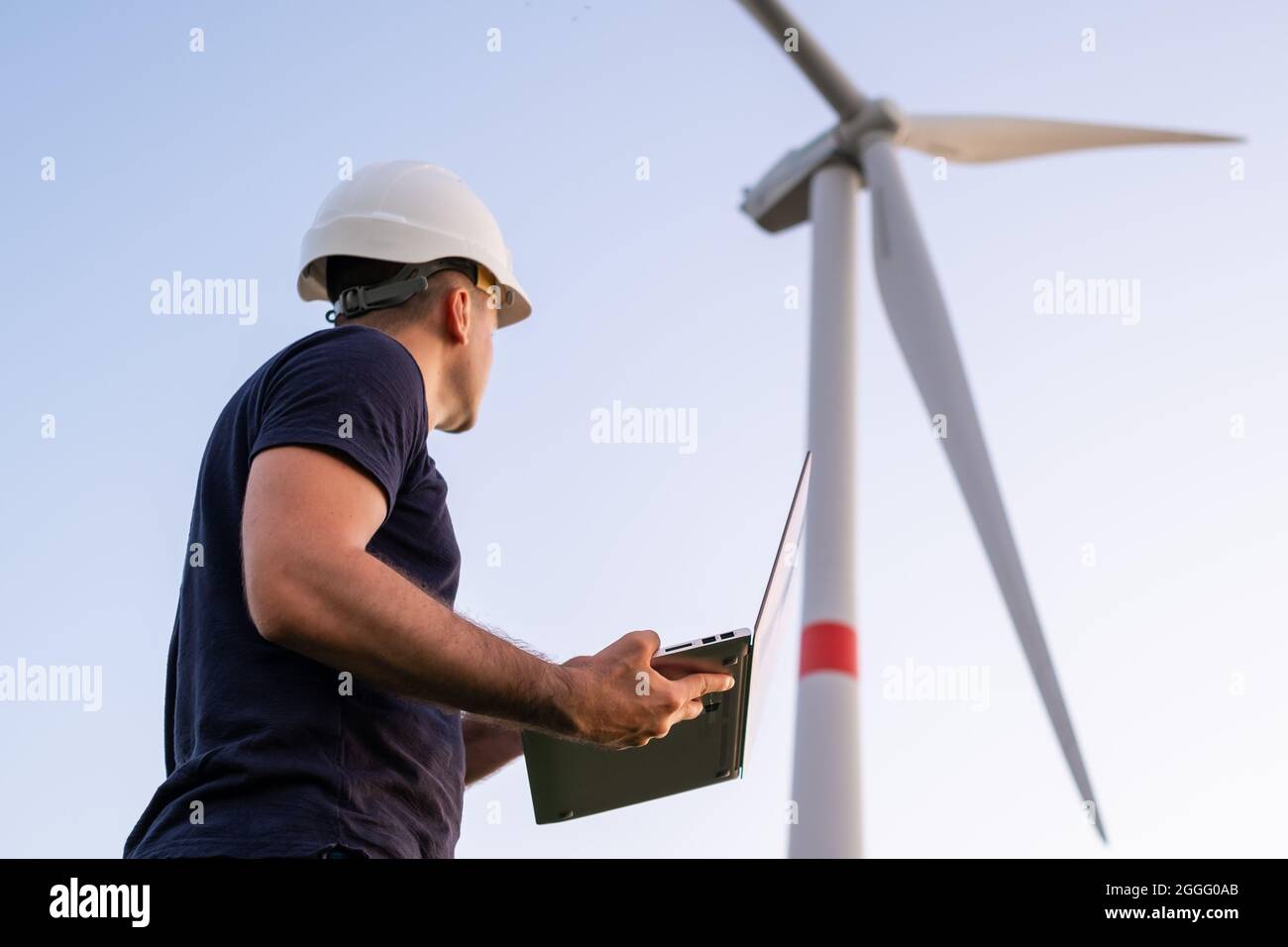 Male engineer in the helmet controls and maintains the operation of a wind turbine using a laptop on the background of windmill Stock Photo